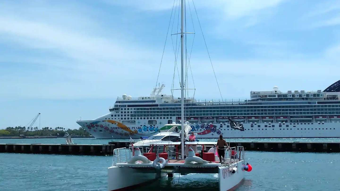 Catamaran approaching dock with a large cruise ship in the background on a sunny day.