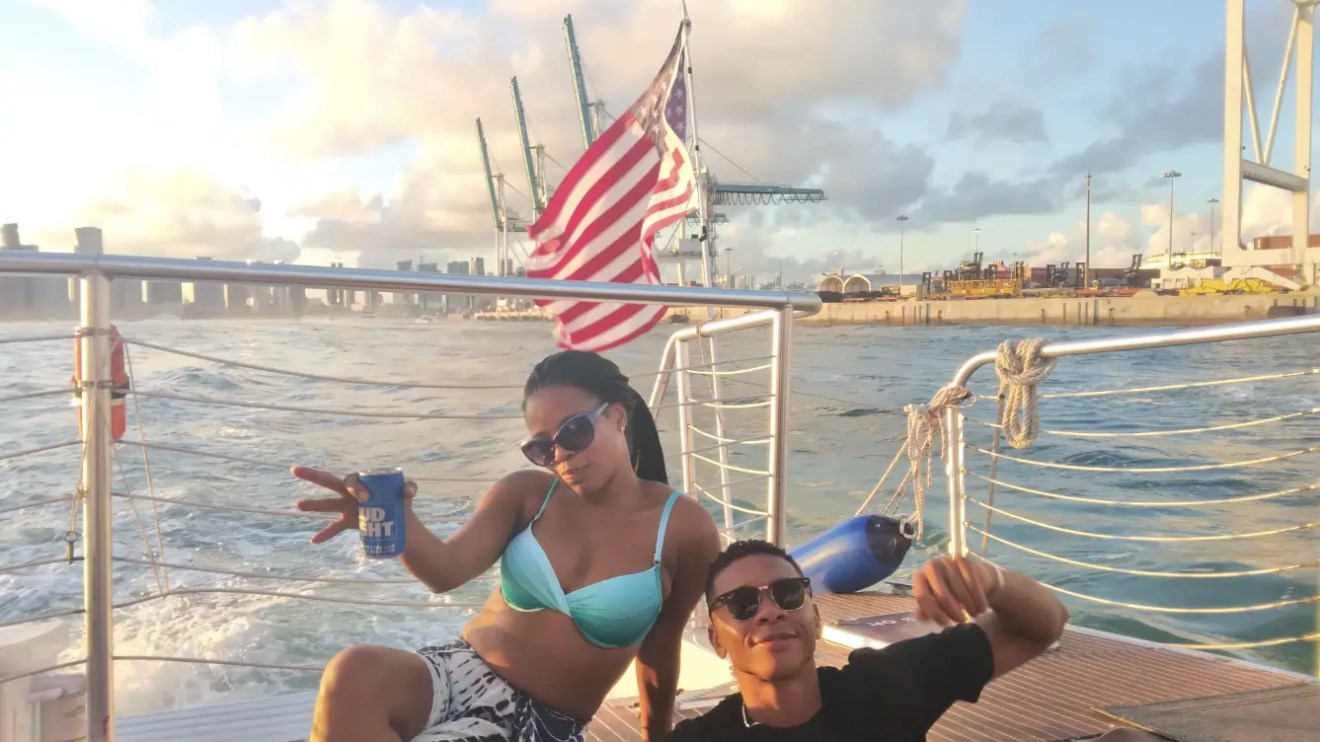 Two people on a boat, one holding a drink, with an American flag waving in the background.