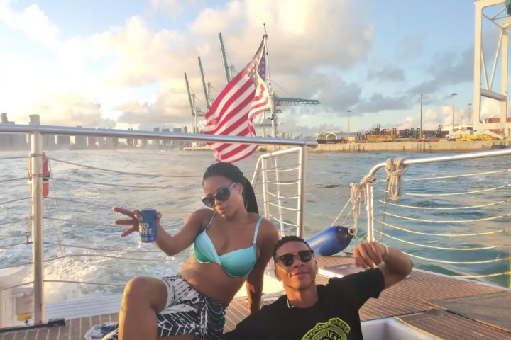Two people on a boat, one holding a drink, with an American flag waving in the background.