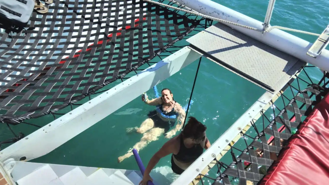Two people swim near a boat with netted decks over the ocean.