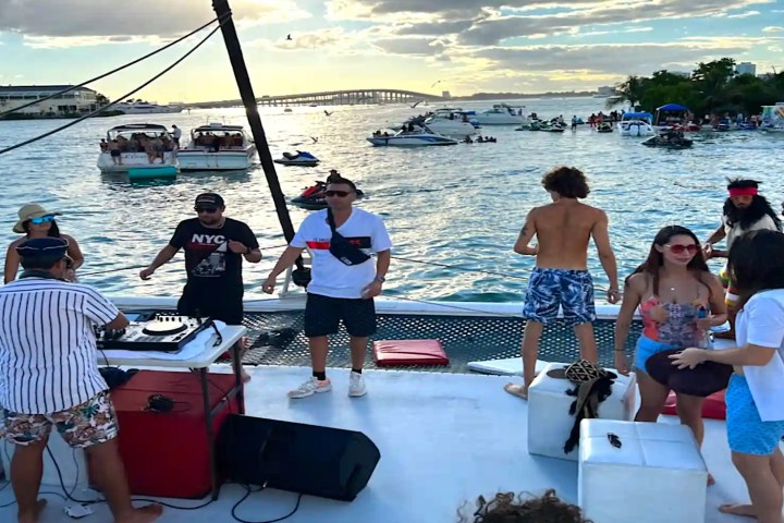 People partying on a boat with a DJ, surrounded by boats on the water at sunset.
