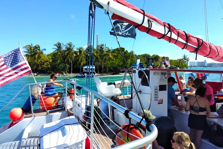 People on a boat with American flag and pirate flag, near tropical shore.