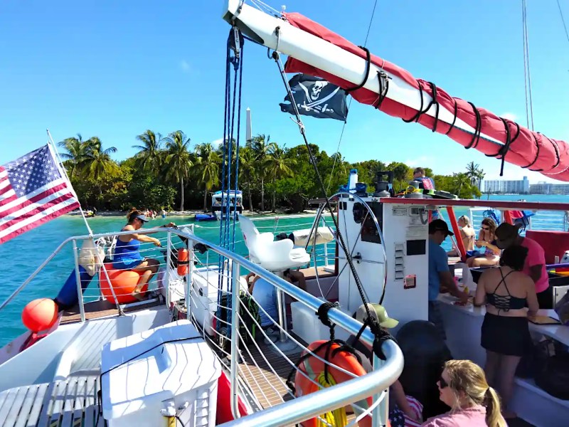 People on a boat with American flag and pirate flag, near tropical shore.