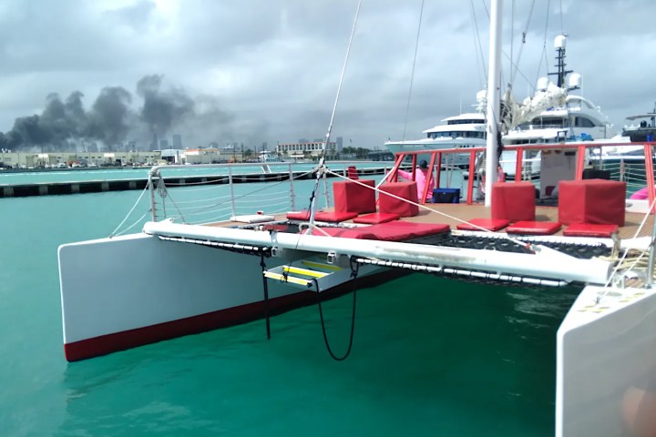Catamaran with red seating docked near yachts, city and smoke in background.