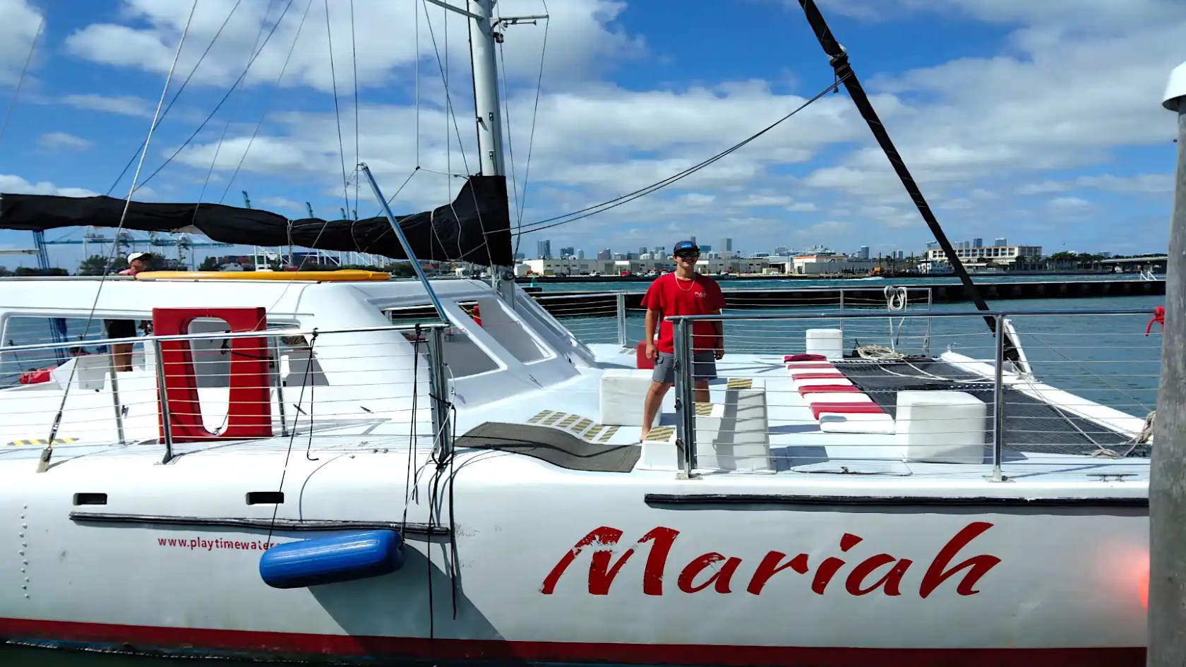 Person in red shirt standing on a sailboat named 'Mariah' docked at a marina.
