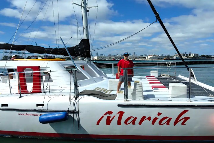 Person in red shirt standing on a sailboat named 'Mariah' docked at a marina.