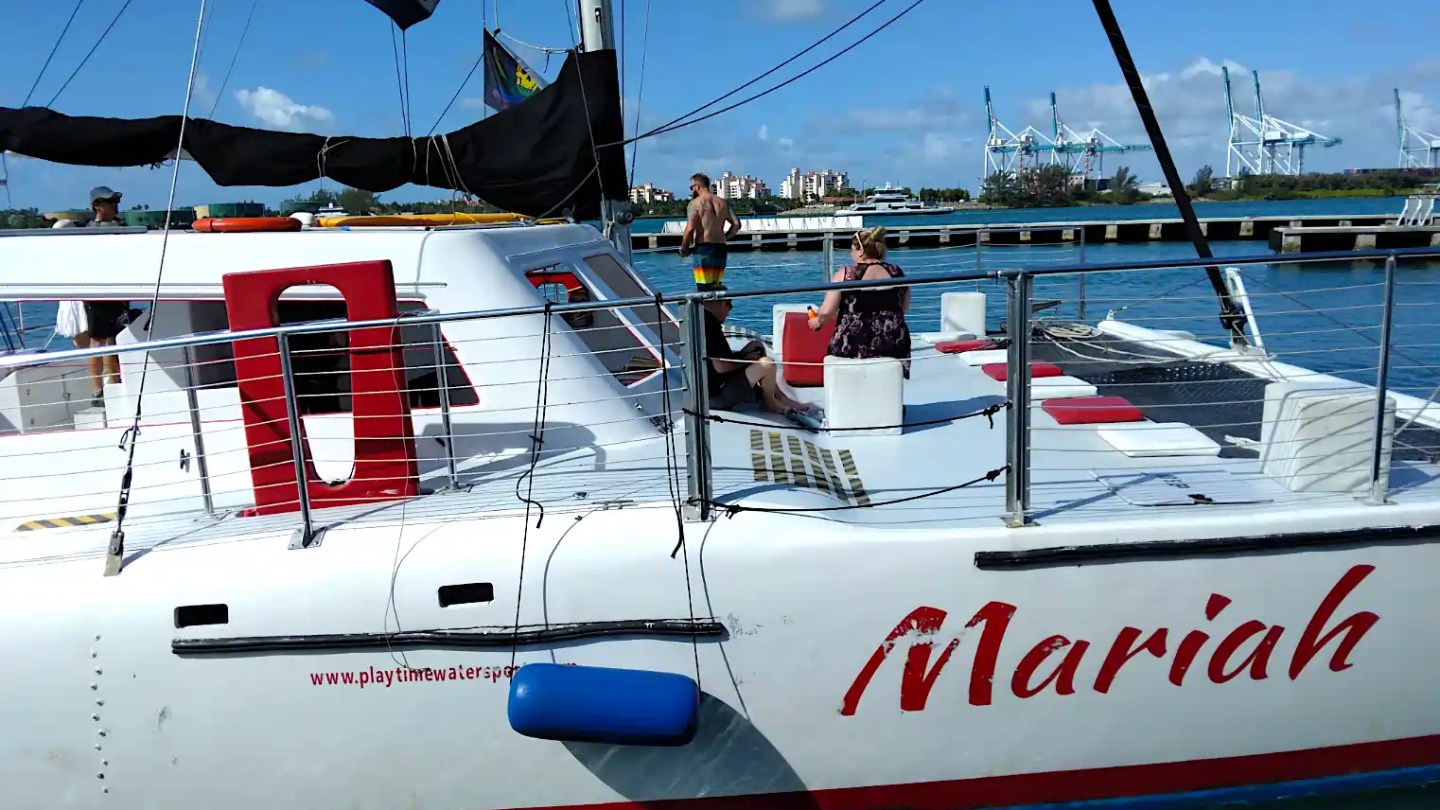 People relaxing on a catamaran named 'Mariah' by a harbor with cranes visible in the background.