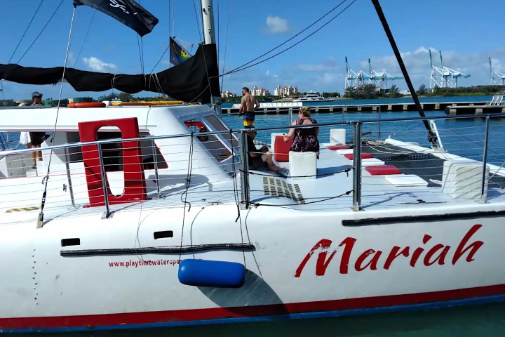 People relaxing on a catamaran named 'Mariah' by a harbor with cranes visible in the background.