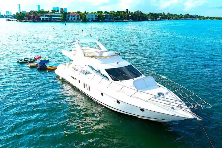 White yacht on blue water with small boats tethered, city skyline in background.