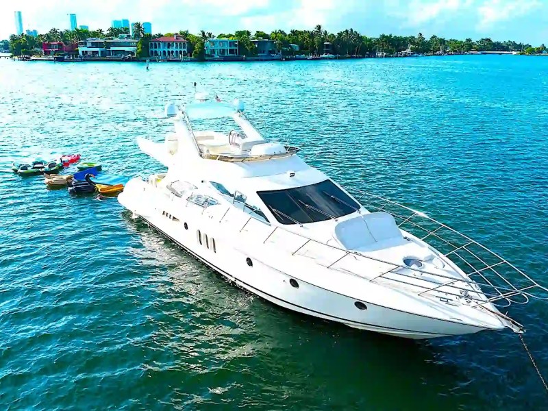 White yacht on blue water with small boats tethered, city skyline in background.