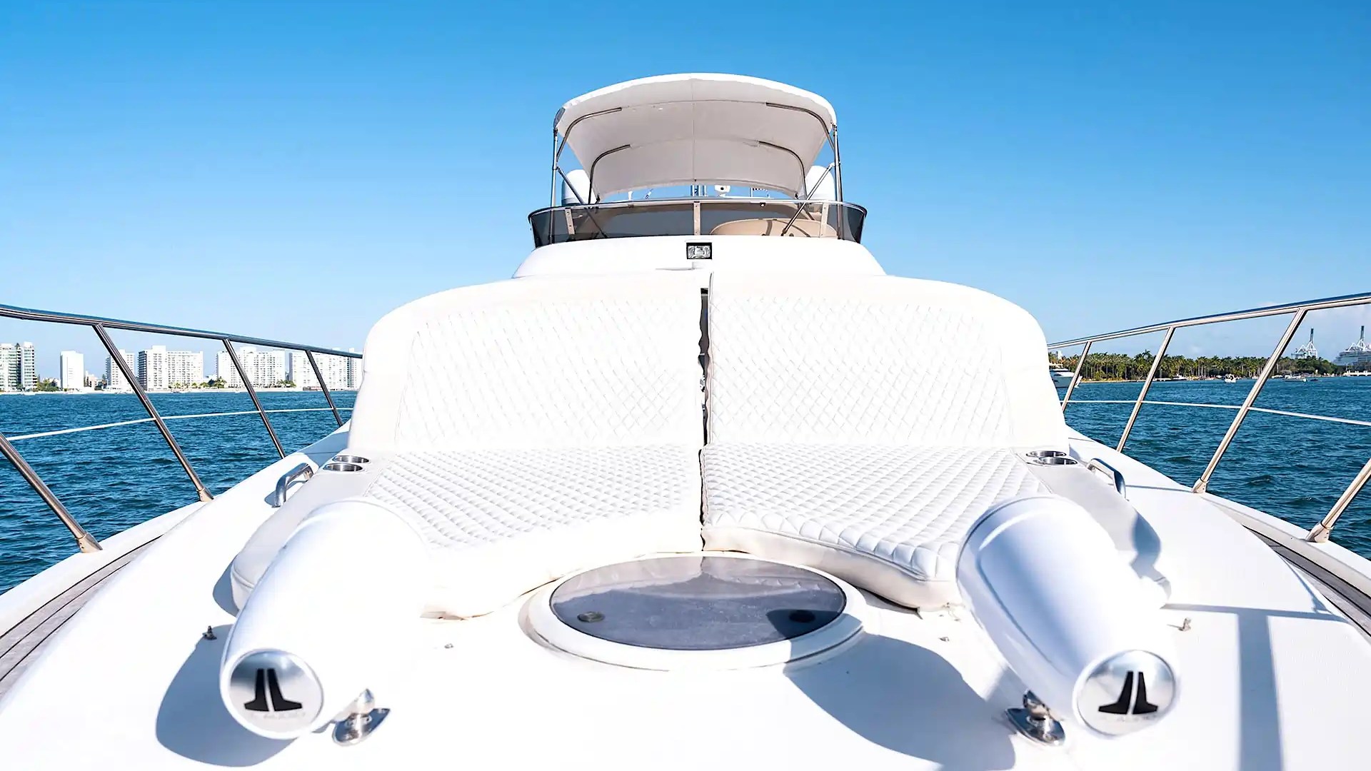 Luxury yacht deck with white lounge seating and speakers, ocean and buildings in background.