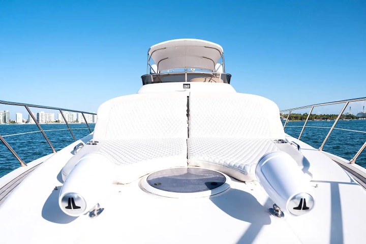 Luxury yacht deck with white lounge seating and speakers, ocean and buildings in background.