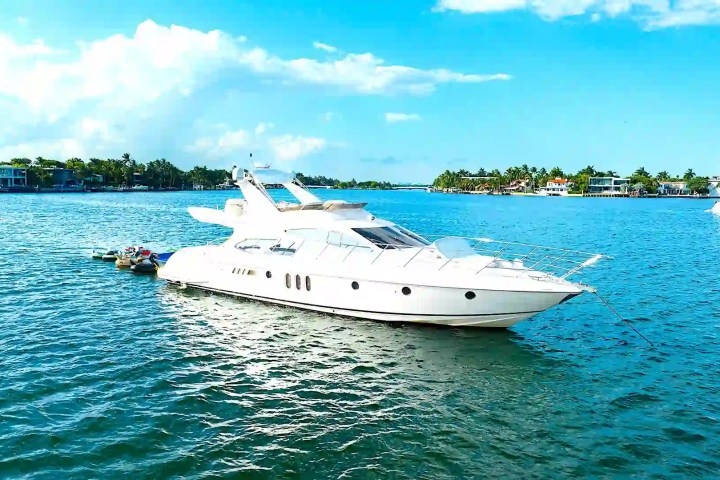 White yacht anchored on blue water with houses and trees in the background.