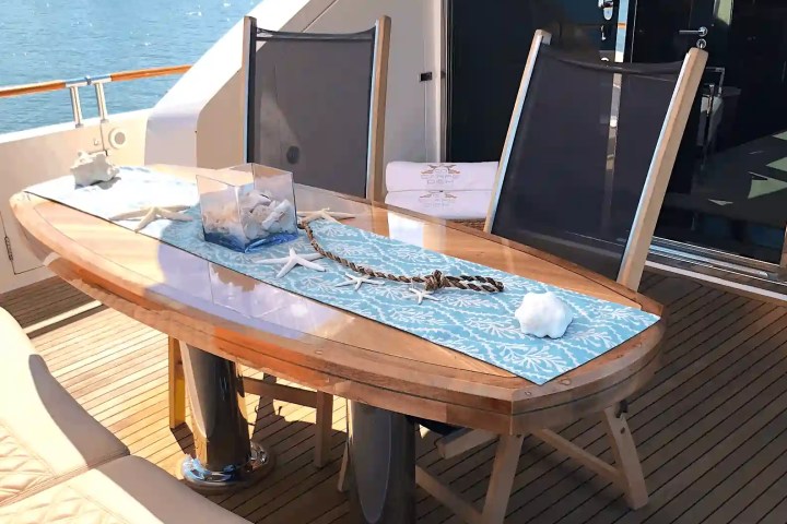 Deck table with blue runner, seashells, and chairs on a yacht with water view.