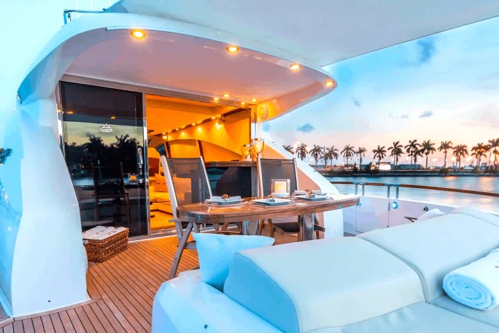 Luxury yacht deck with dining table, lounge chair, and palm trees by a sunset-lit shore.
