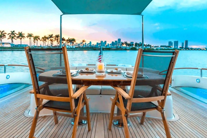 Yacht deck with dining table, two chairs, city skyline, and palm trees at sunset.