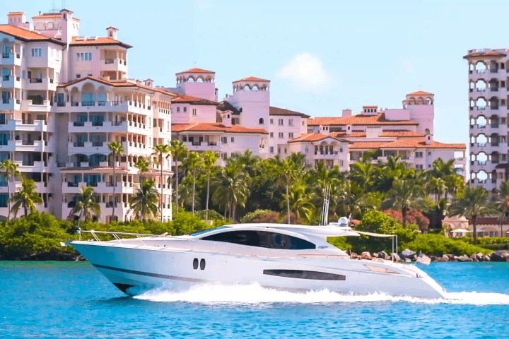 A sleek white yacht cruising on blue water with luxury buildings and palm trees in the background.