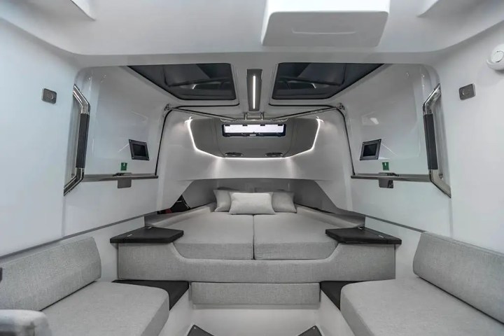 Modern boat interior with gray seating and a central bed, featuring sleek lines and windows overhead.