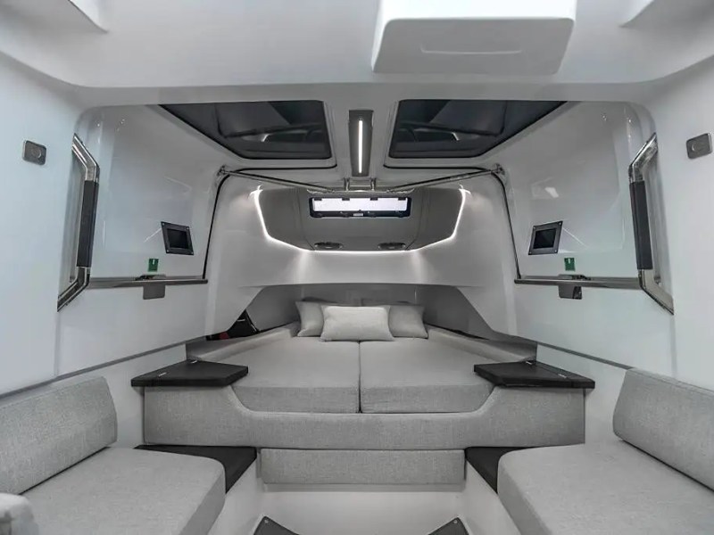 Modern boat interior with gray seating and a central bed, featuring sleek lines and windows overhead.