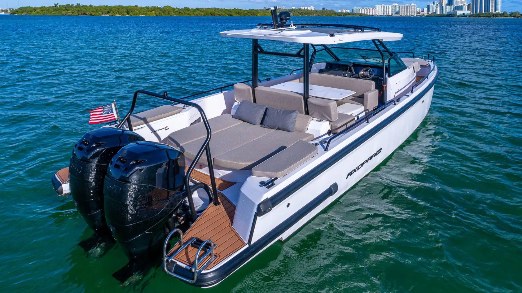 White motorboat with dual engines cruising on blue water, city skyline in background.