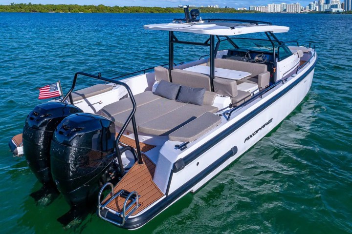White motorboat with dual engines cruising on blue water, city skyline in background.
