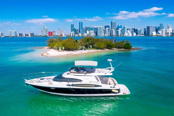Luxury yacht near a small island with city skyline in the background under a clear blue sky.