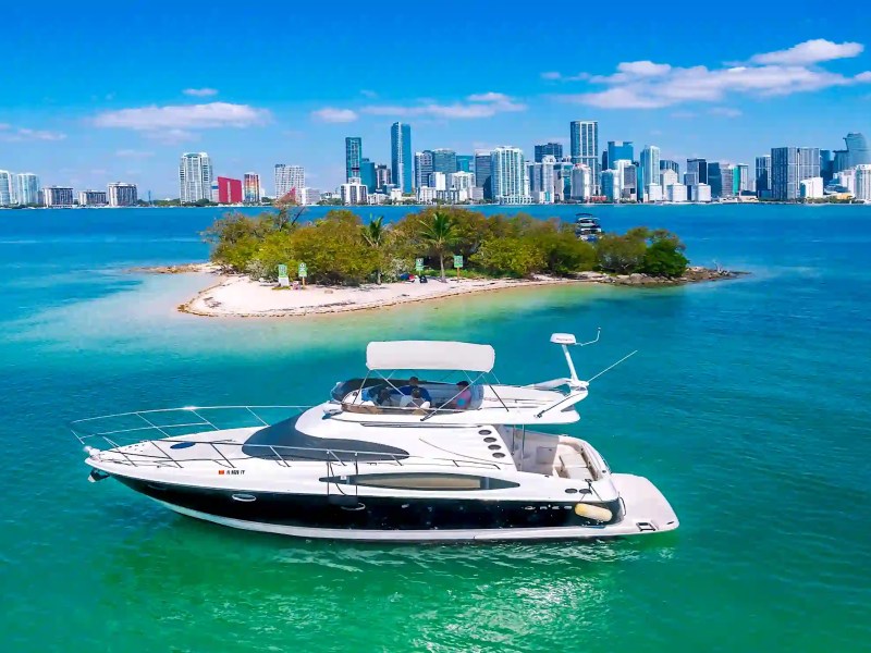 Luxury yacht near a small island with city skyline in the background under a clear blue sky.