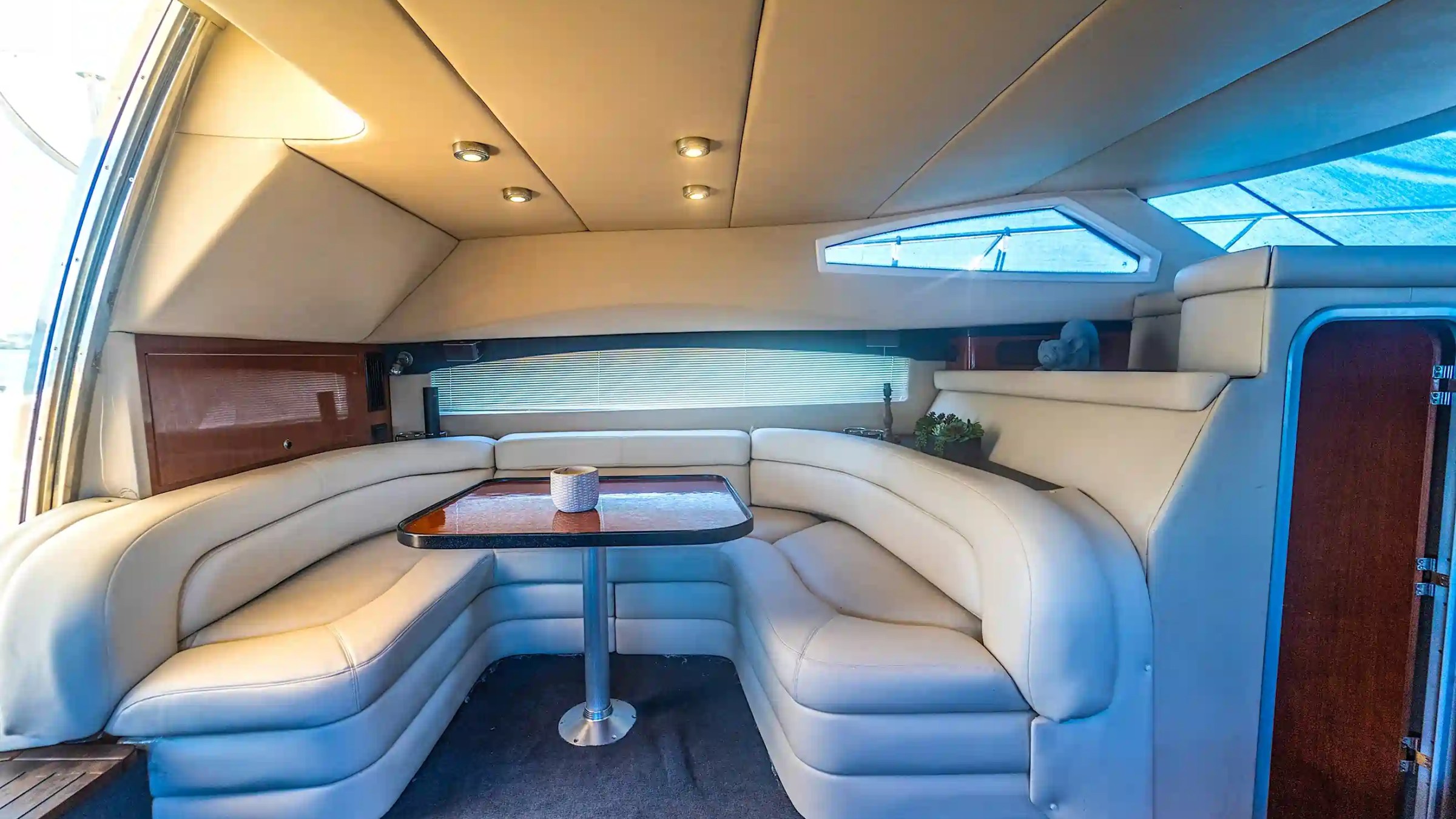 Interior of a yacht with a curved white leather sofa around a wooden table and recessed lighting.