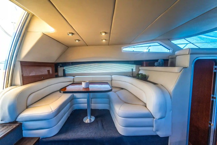 Interior of a yacht with a curved white leather sofa around a wooden table and recessed lighting.