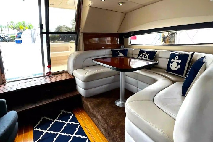 Yacht interior with white L-shaped sofa, nautical pillows, and wooden table.