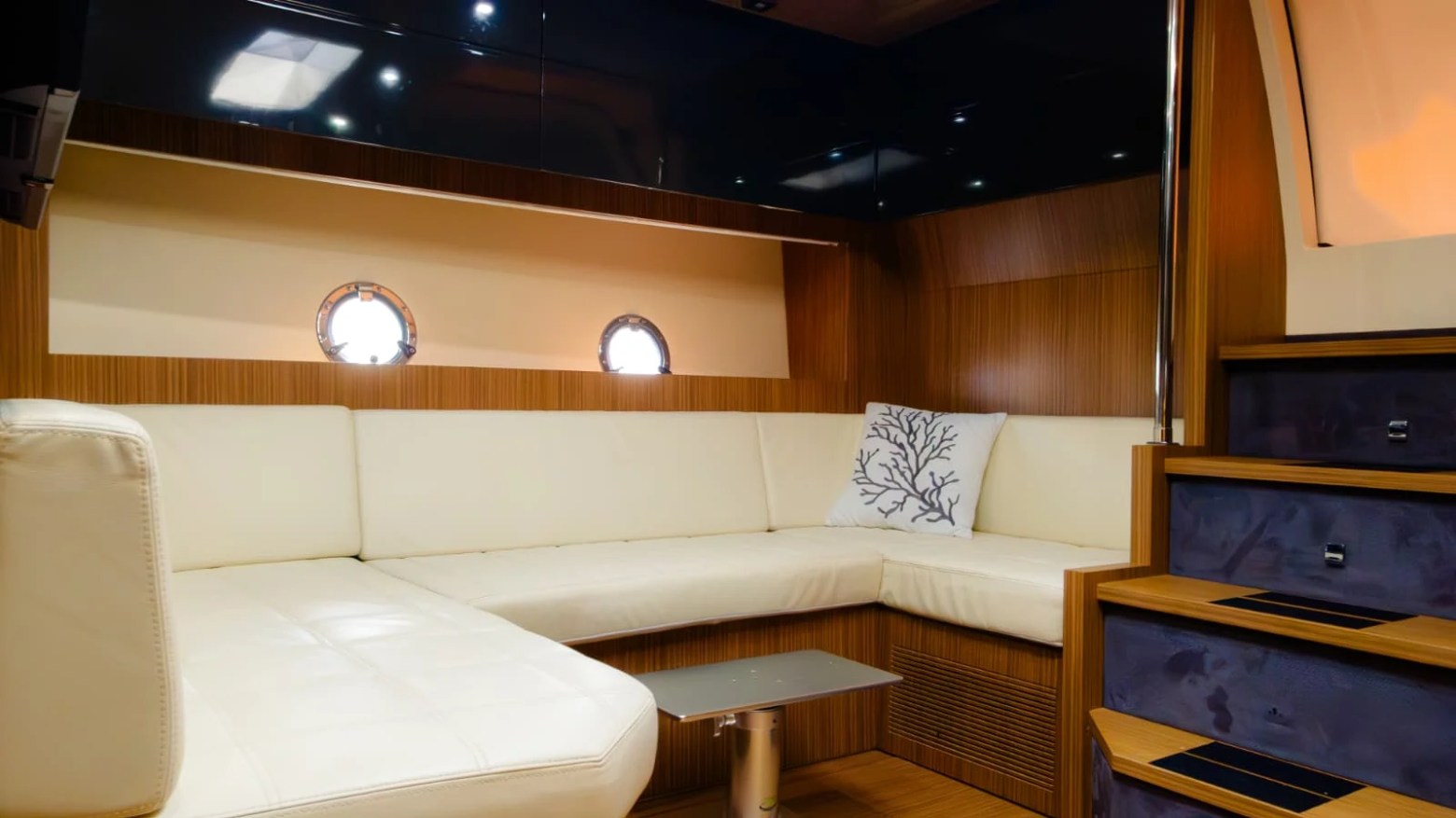 Luxury yacht interior with white L-shaped sofa, wood paneling, portholes, and stairs.