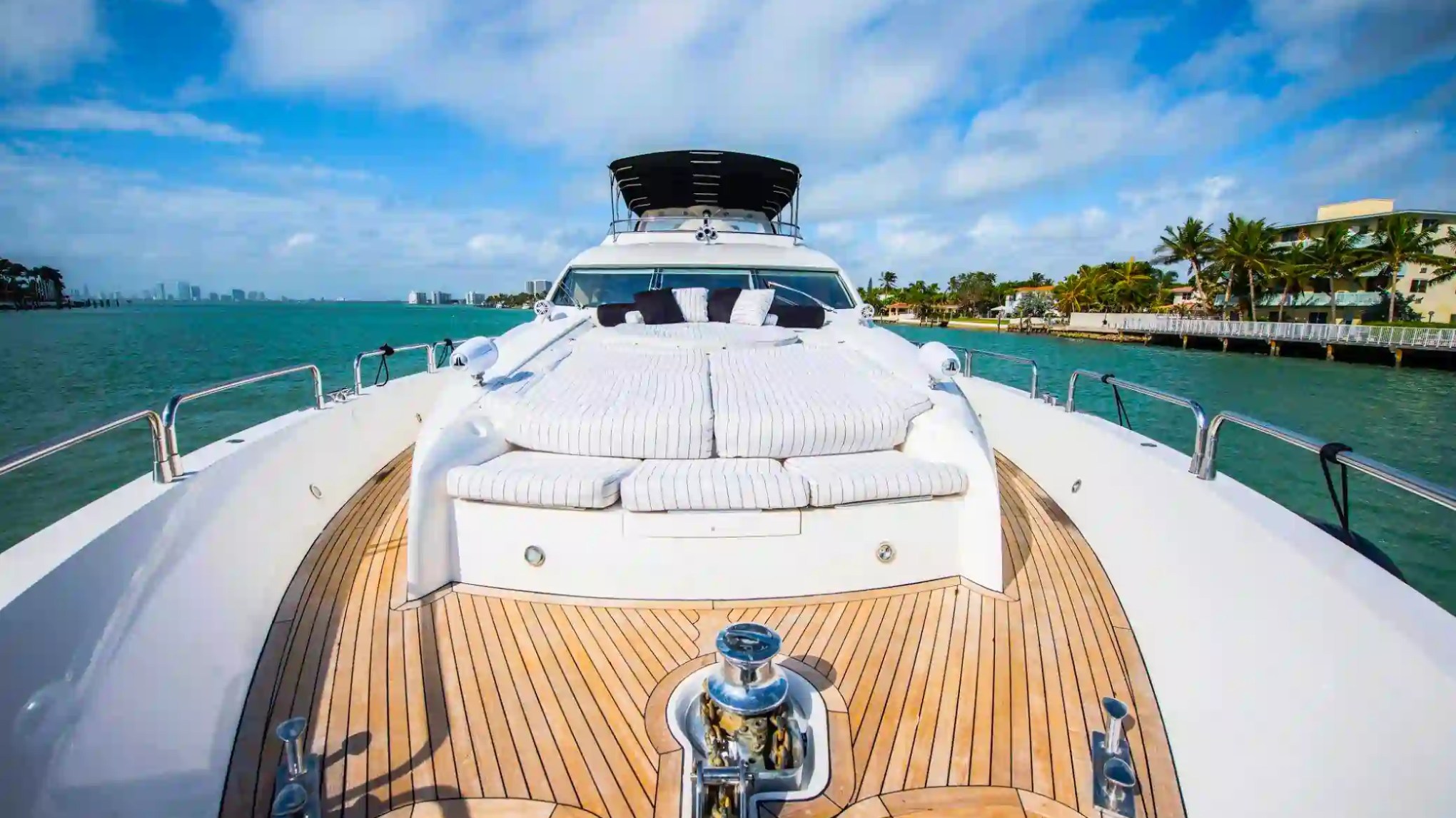 Luxury yacht deck with sunbeds, surrounded by water, palm trees, and a city skyline under a cloudy blue sky.