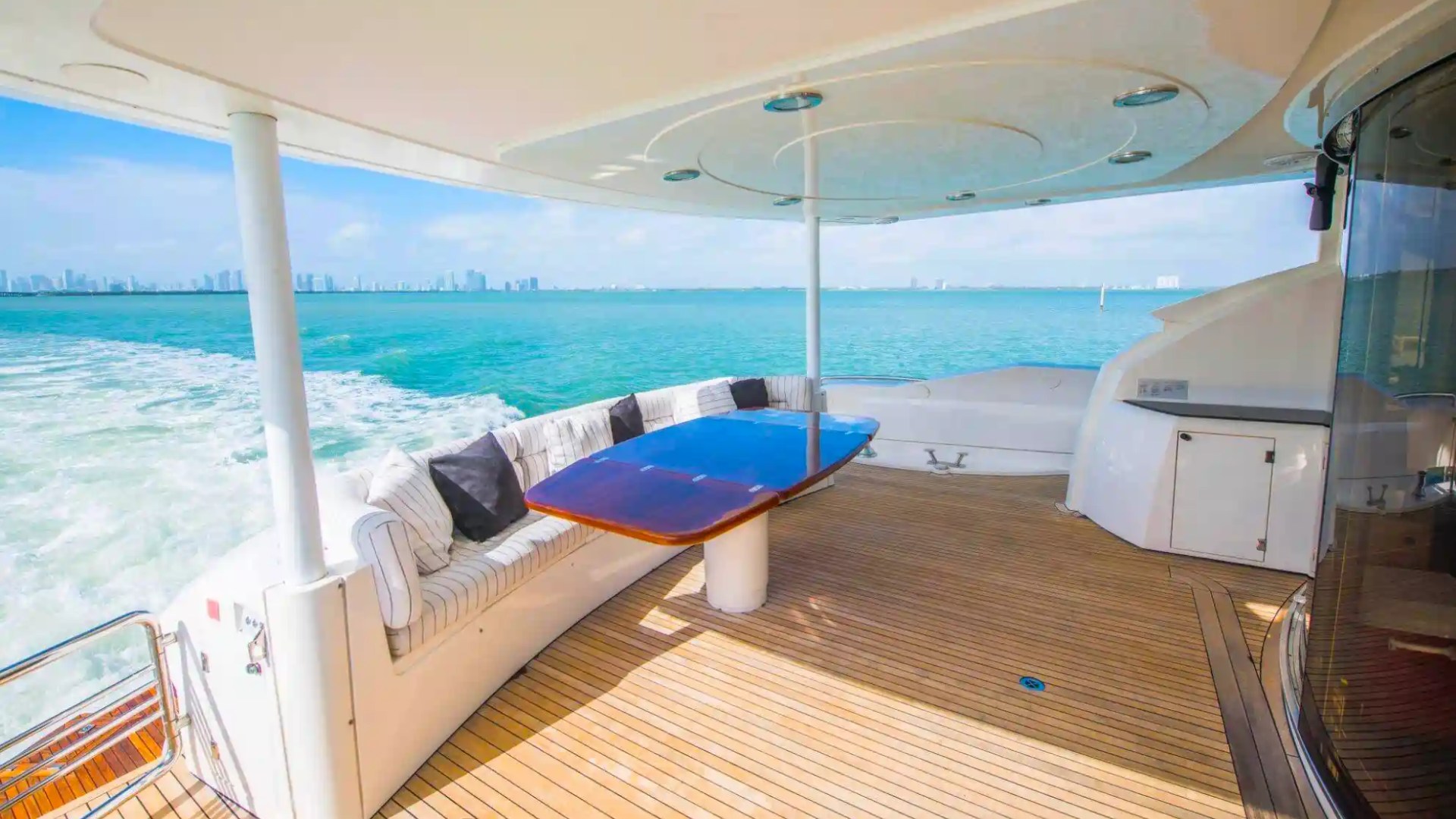 Yacht deck with table and seating, ocean view, city skyline in distance.