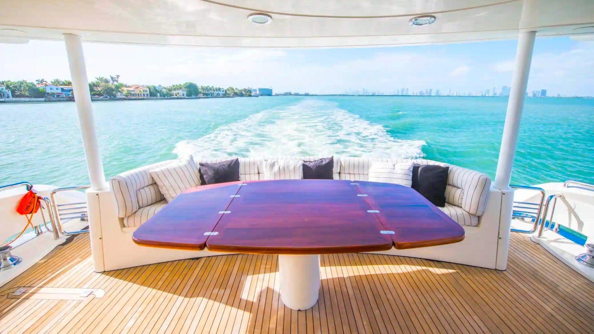 Rear deck of a yacht with a table and seating area overlooking the ocean and distant city skyline.