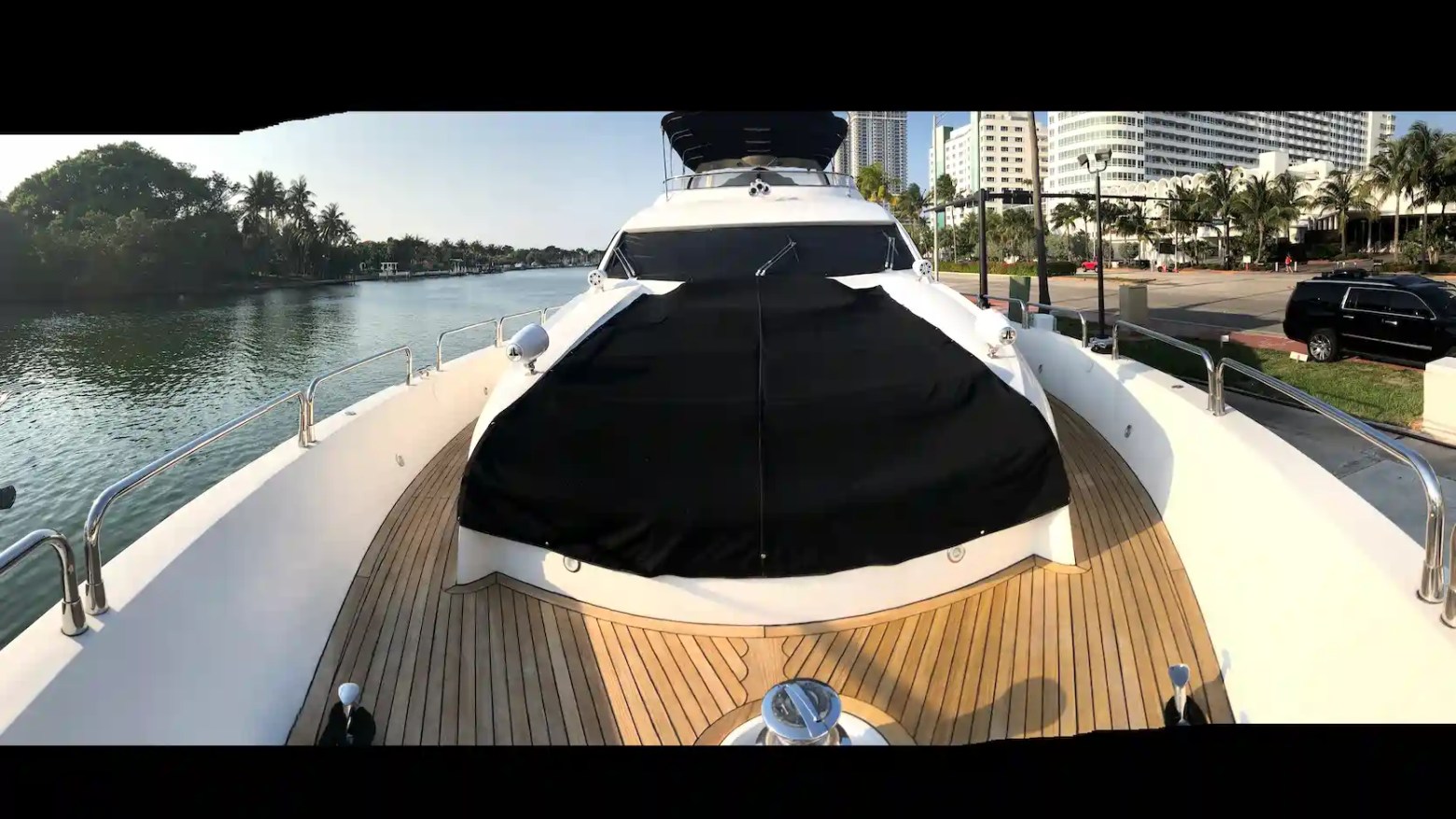 Bow of a yacht with wooden deck, docked near water, city buildings and trees in the background.