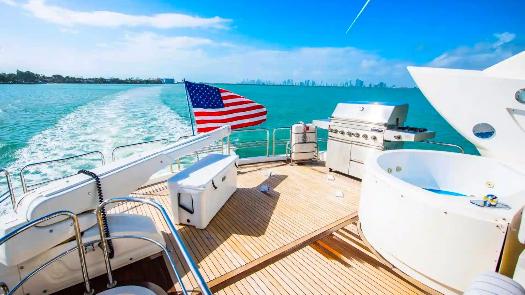 Yacht deck with American flag, jacuzzi, grill, and ocean view, clear blue skies.
