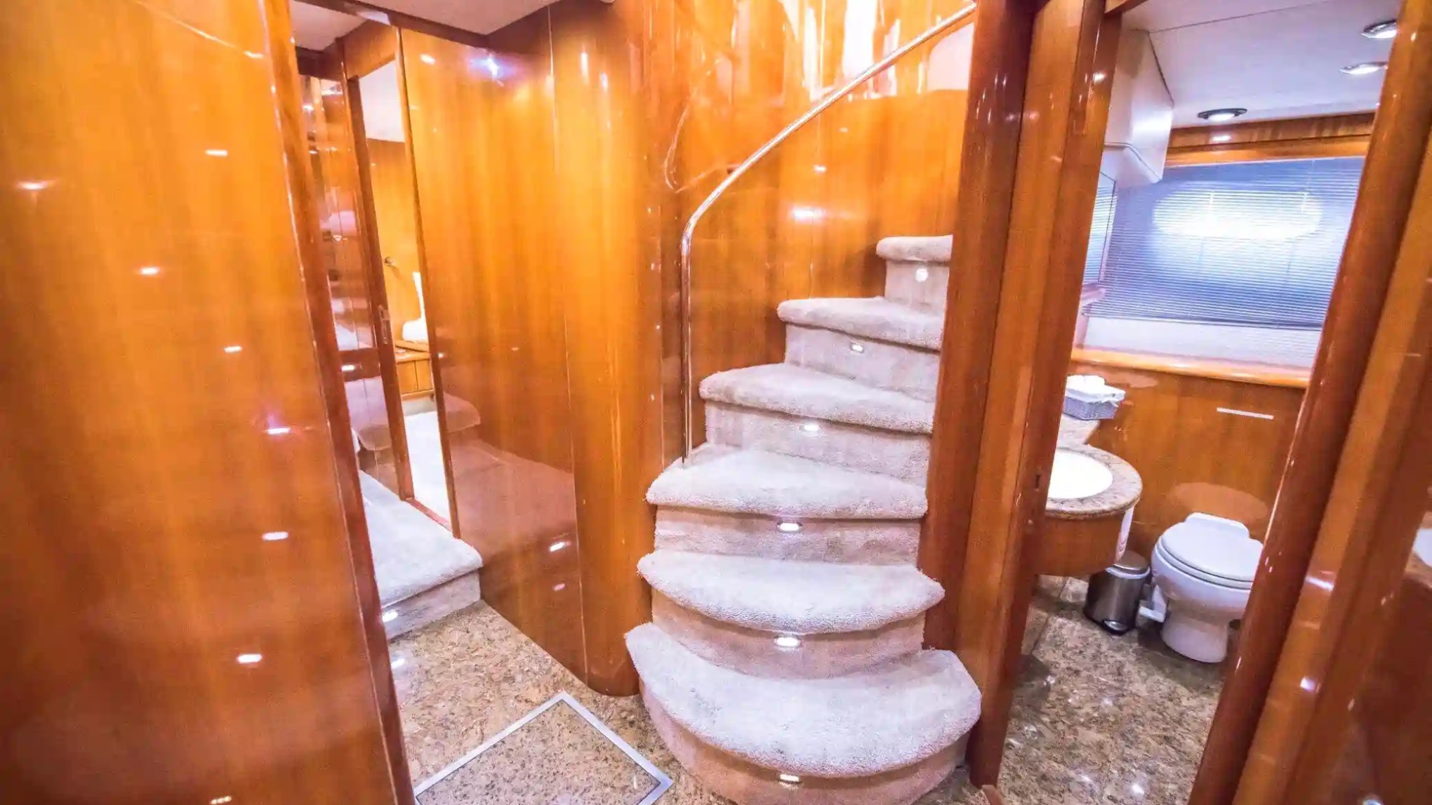 Elegant yacht interior with curved stairs, glossy wood paneling, and a bathroom with marble floor and toilet.
