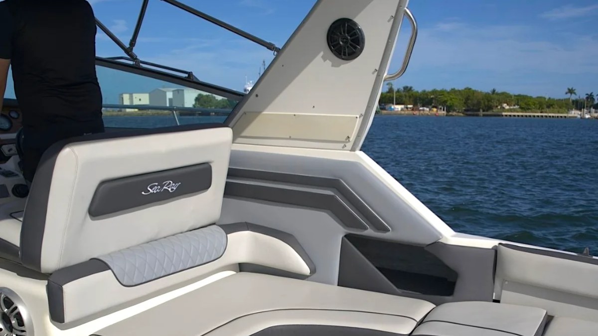 Person steering a boat with gray seating in open water, bright sky.