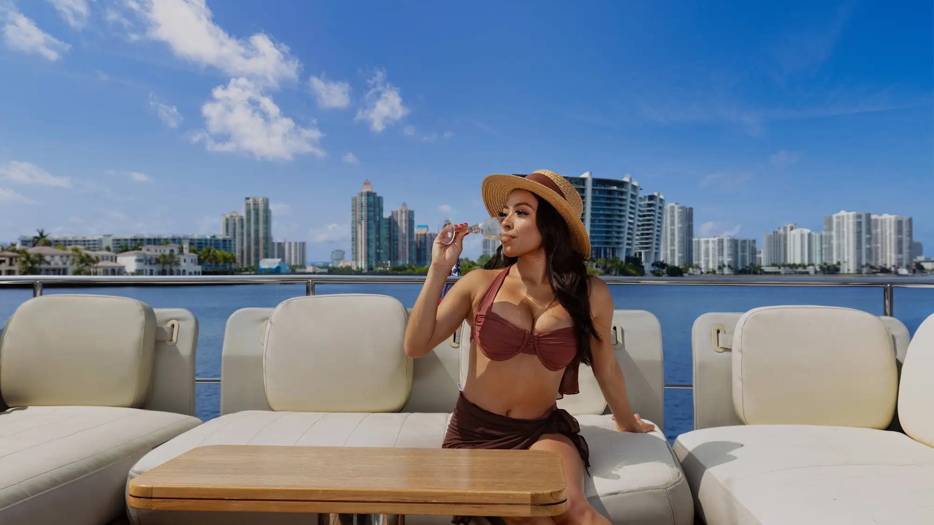 Woman in a hat sits on a boat with city skyline in background.