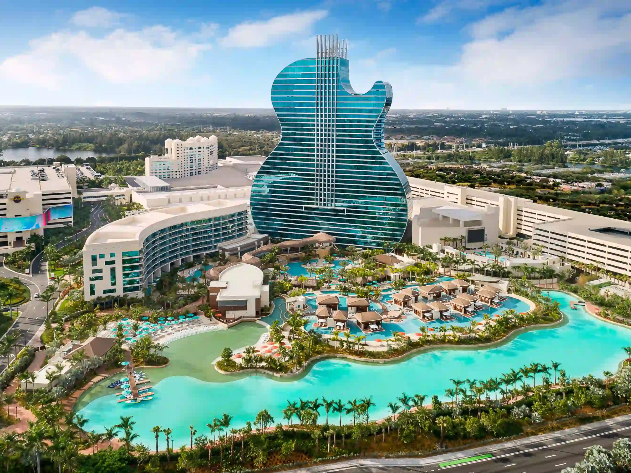Aerial view of a guitar-shaped hotel and resort with pools and palm trees.