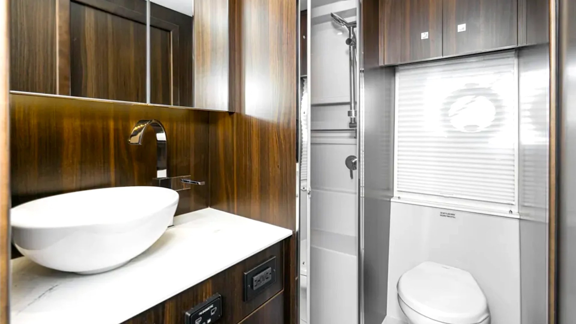 Modern RV bathroom with wooden panels, white sink, toilet, and shower.