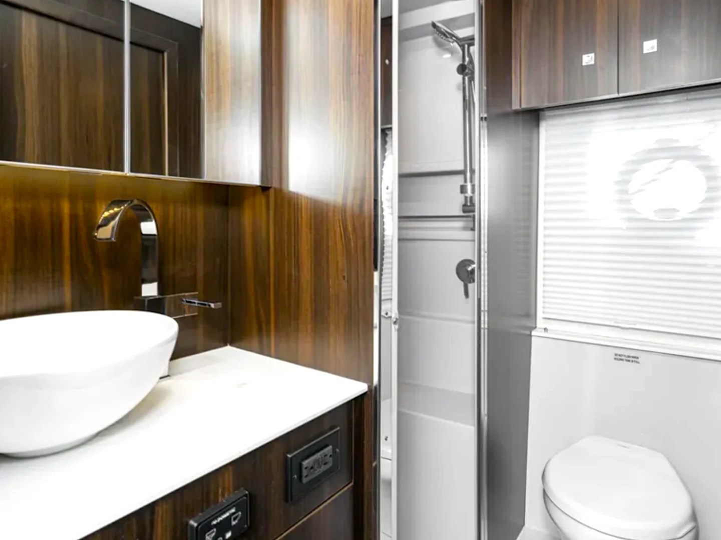 Modern RV bathroom with wooden panels, white sink, toilet, and shower.