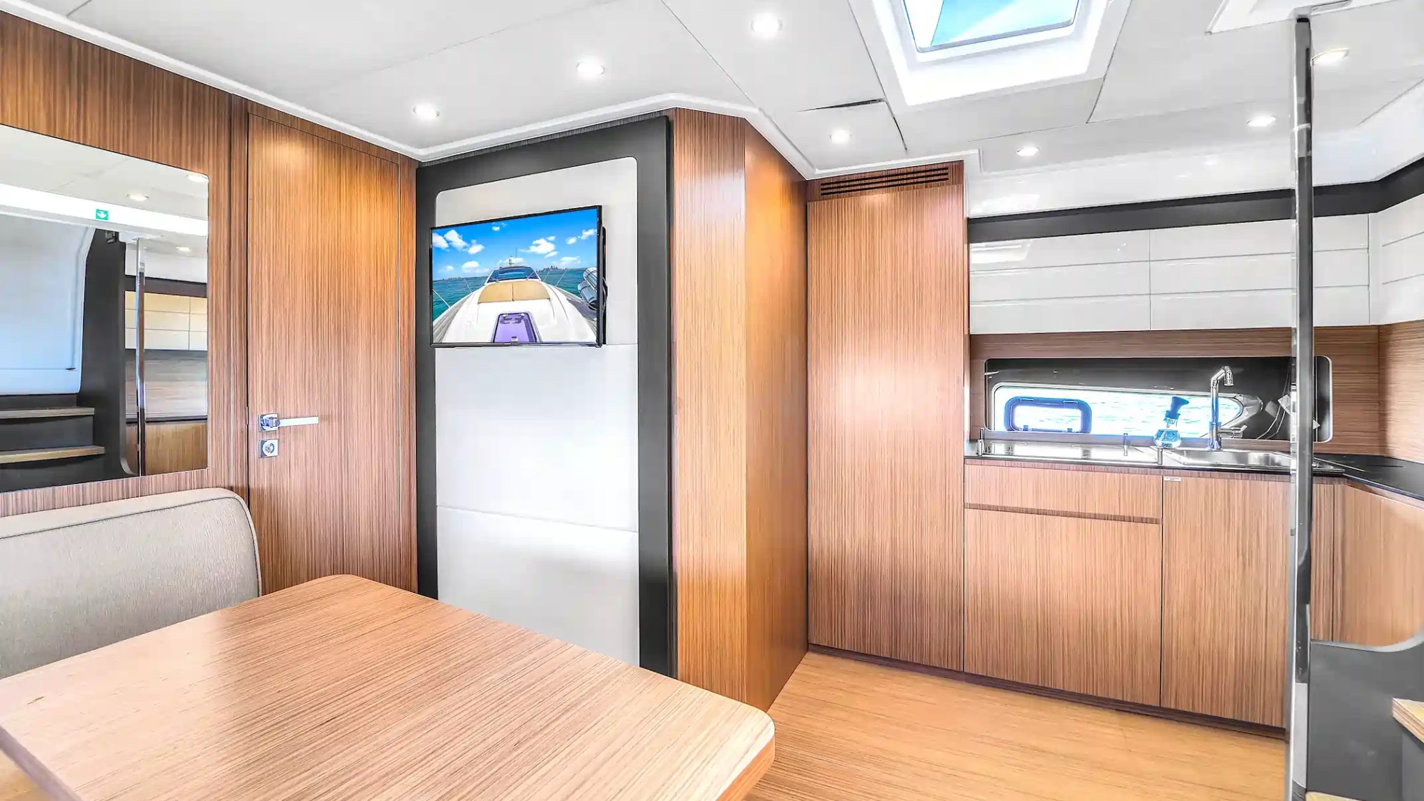 Modern yacht interior with wood paneling, table, TV, skylight, and kitchenette.