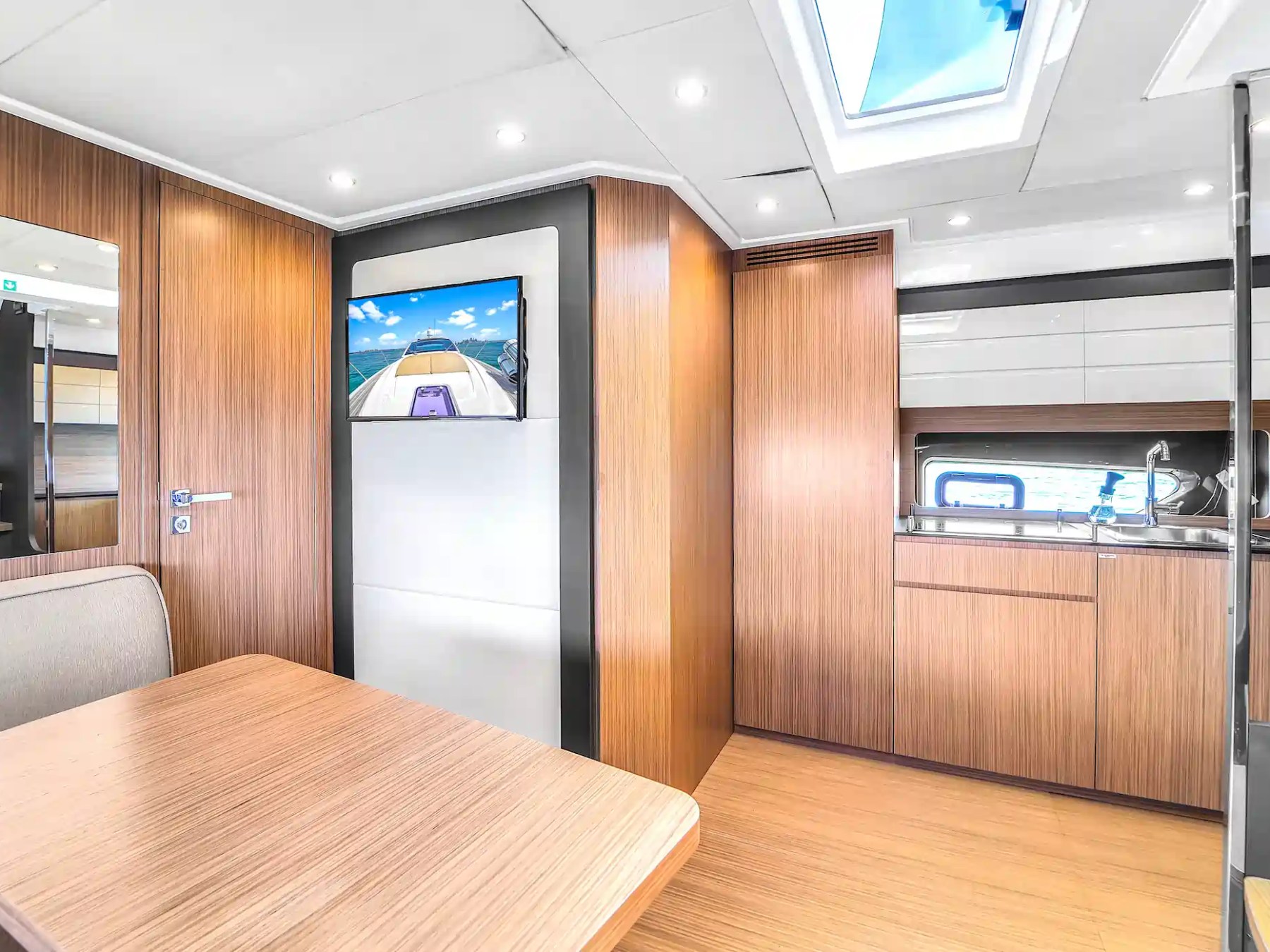 Modern yacht interior with wood paneling, table, TV, skylight, and kitchenette.