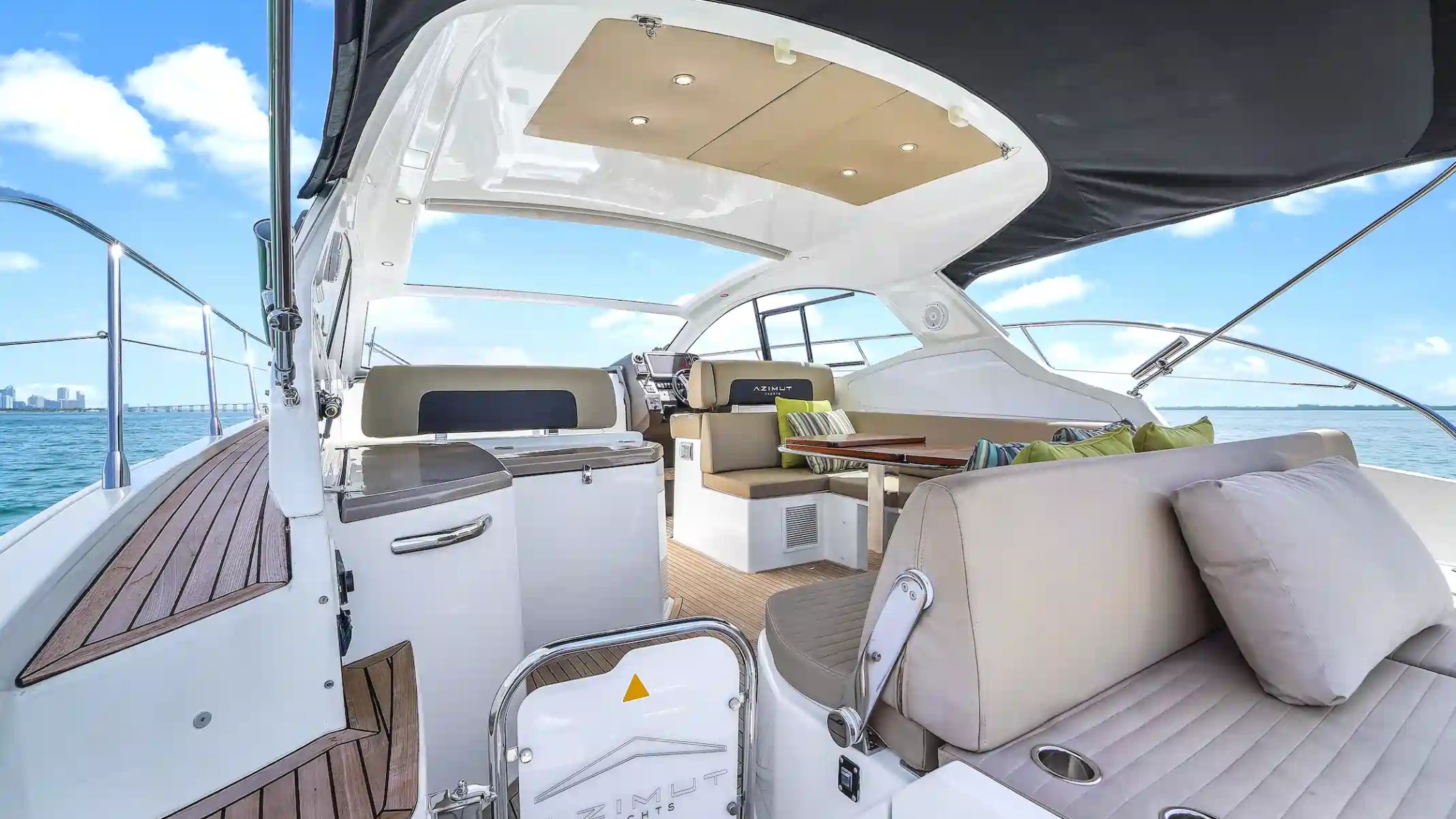 Luxury yacht deck with seating area, cushions, and table under a canopy on a sunny day.