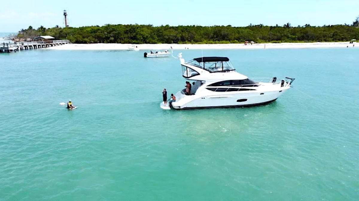This 44 foot Meridian Boat Rental is anchored in crystal-clear turquoise waters near Cape Coral with guests enjoying the swim platform while a sandy island shoreline and lighthouse are visible in the background.