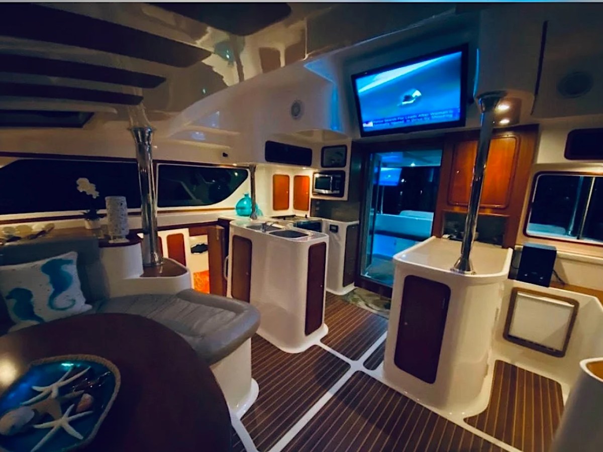 This 50 foot AfriCat Yacht Charter showcases an illuminated luxury salon with dining seating, galley counters, entertainment screen, and ambient evening lighting while cruising near Fort Myers and Sanibel Island.