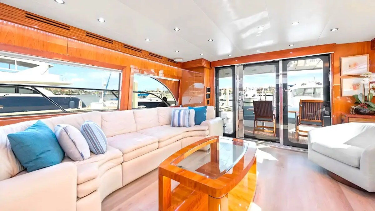 64' Hatteras Yacht Charter salon featuring plush sectional seating, glossy wood finishes, panoramic windows, and seamless access to the aft deck marina views in Marco Island.