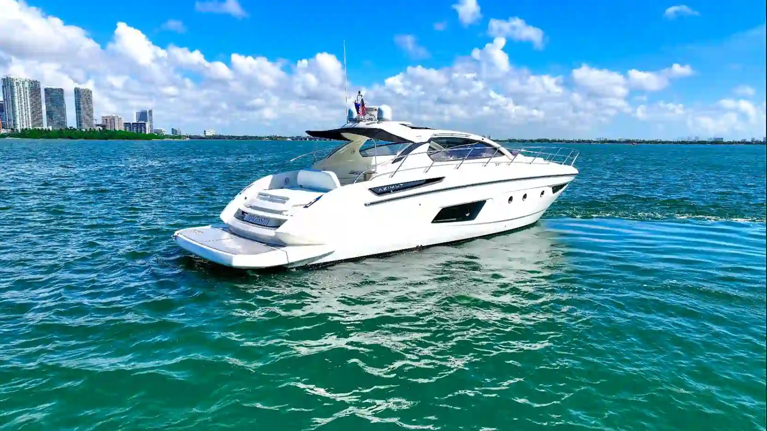 Scenic profile view of a 50' Azimut Yacht Charter floating on Biscayne Bay with Miami skyline buildings, open water, swim platform, and sleek sport yacht design.