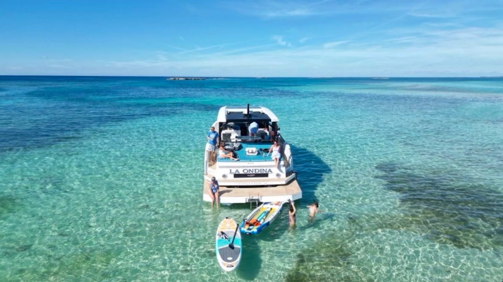 50' DeAntonio Yacht Charter viewed from above in Palm Beach with guests relaxing on the swim platform while paddleboards float in shallow Atlantic Ocean turquoise water.